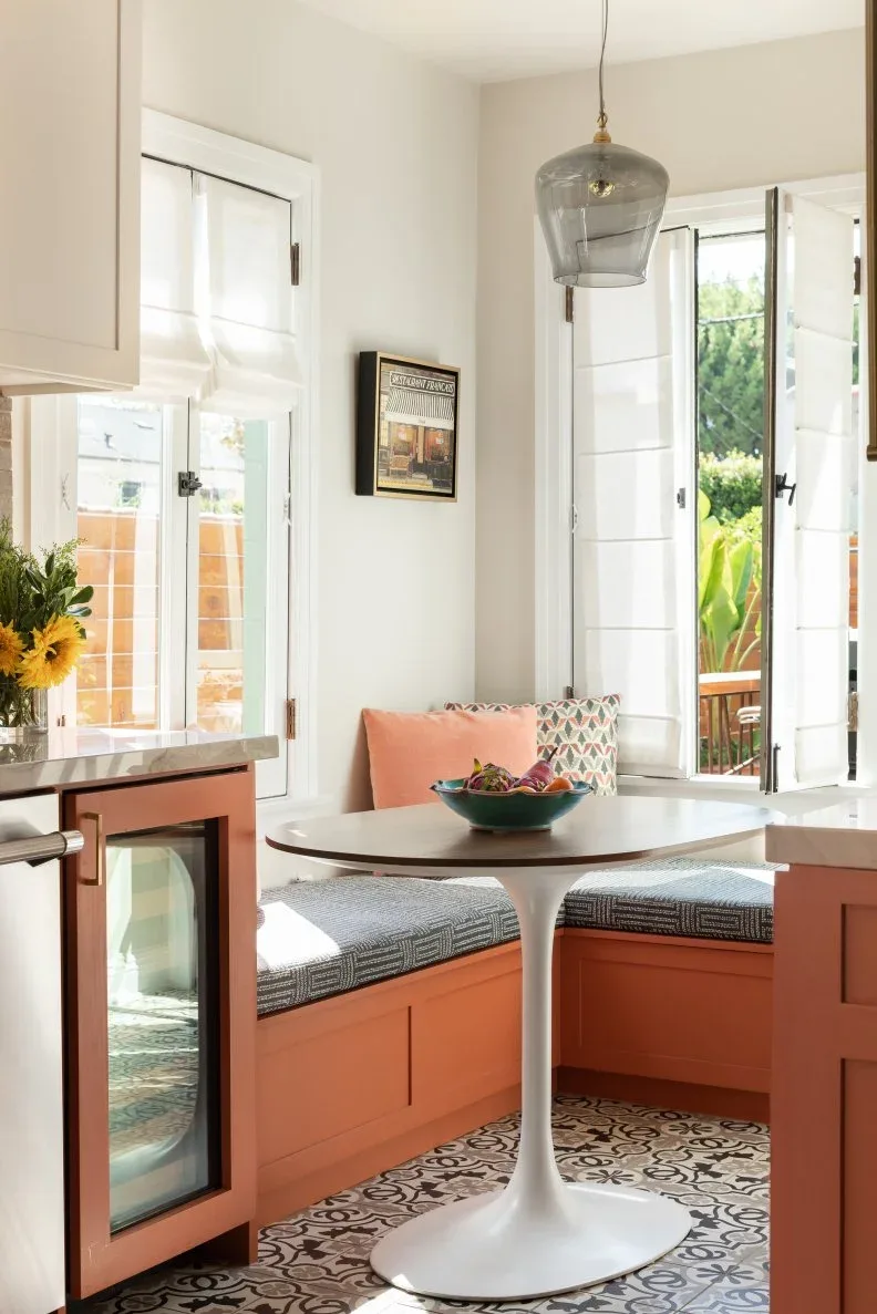 Orange corner banquette with gray patterned seat cushions, a white pedestal table, pale walls, a glass pendant, and windows beside kitchen cabinets.