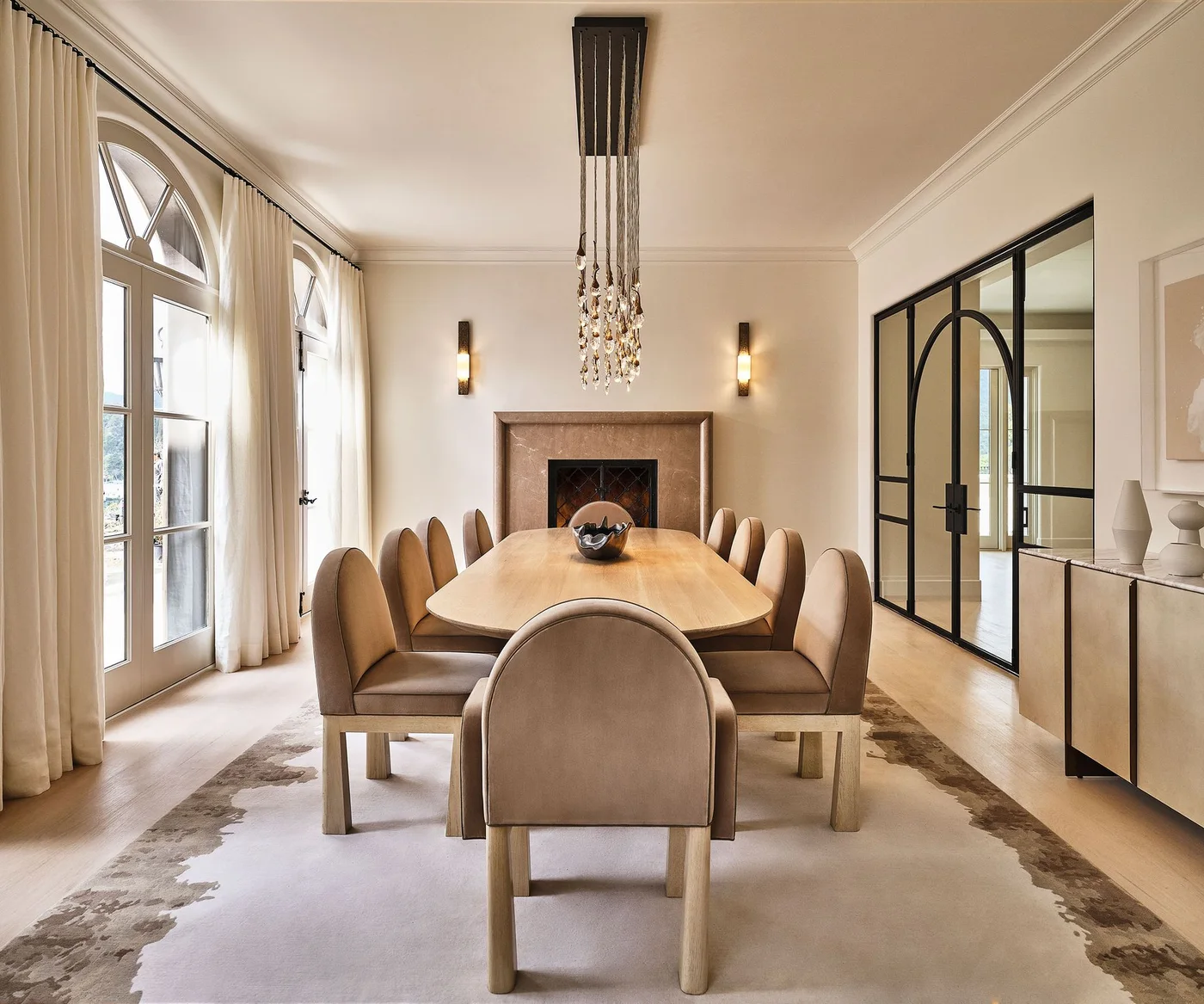 Beige upholstered dining chairs surround a pale wooden table beneath a hanging light between arched windows and black-framed doors