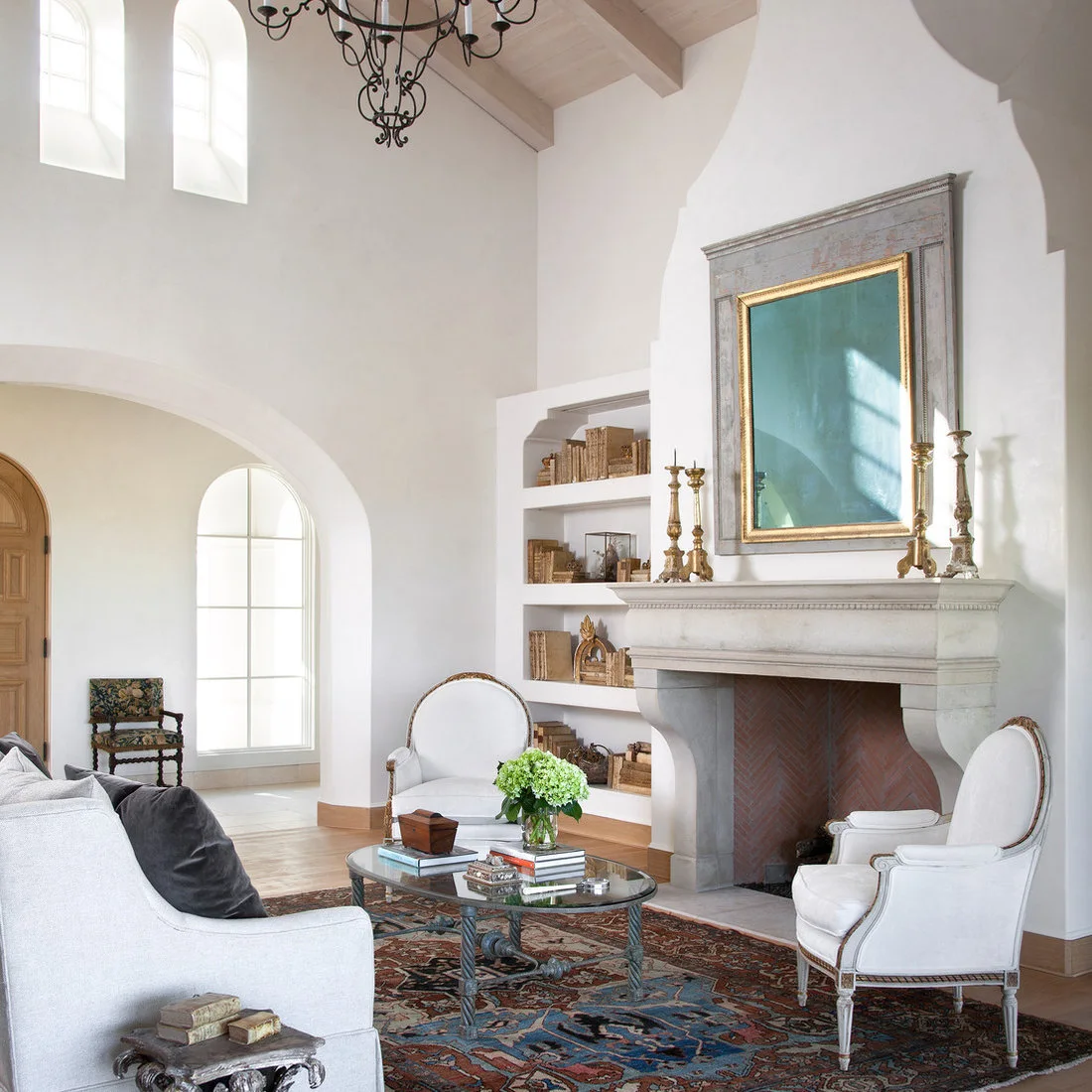A tall pale living room combines arched openings, a curved fireplace outline, white chairs, a pale sofa and patterned rug beneath a decorative chandelier
