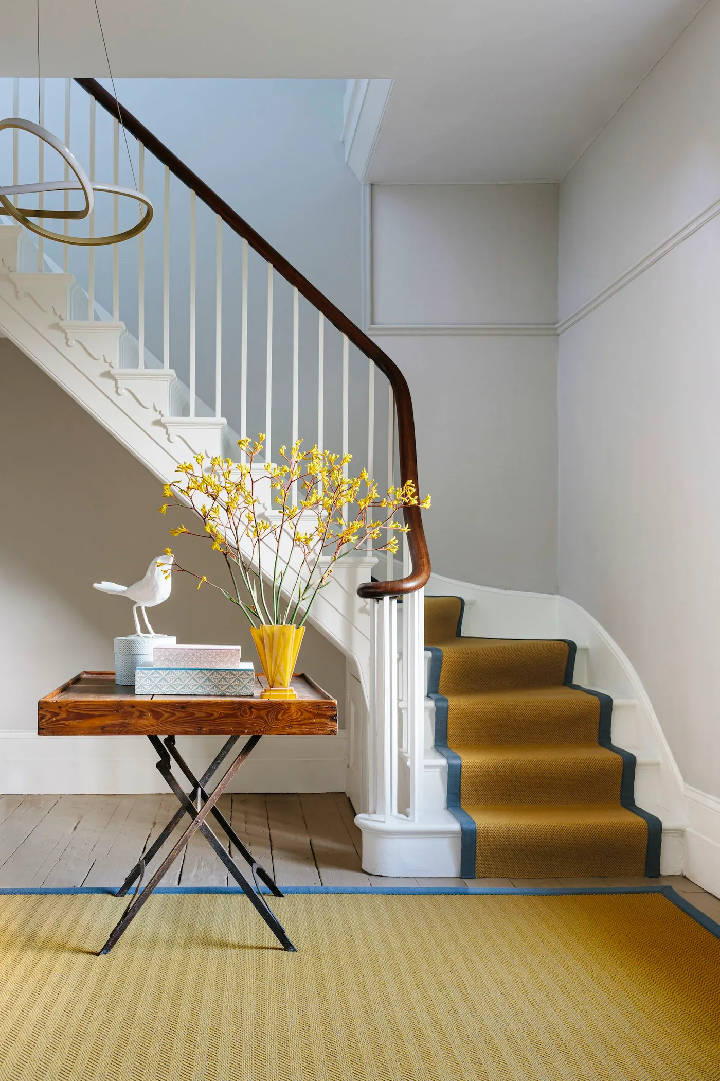Mustard-toned hall rug with blue edging beside pale floorboards, a wood-topped table and a matching stair runner