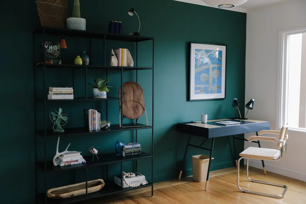 A deep teal-green wall sits behind black open shelves and a dark desk beside a pale woven-seat chair