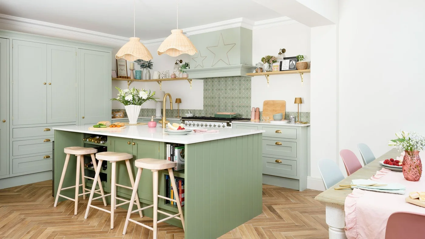 A green island with open shelving sits before pale green tall cabinets and lighter counters beneath woven pendant shades