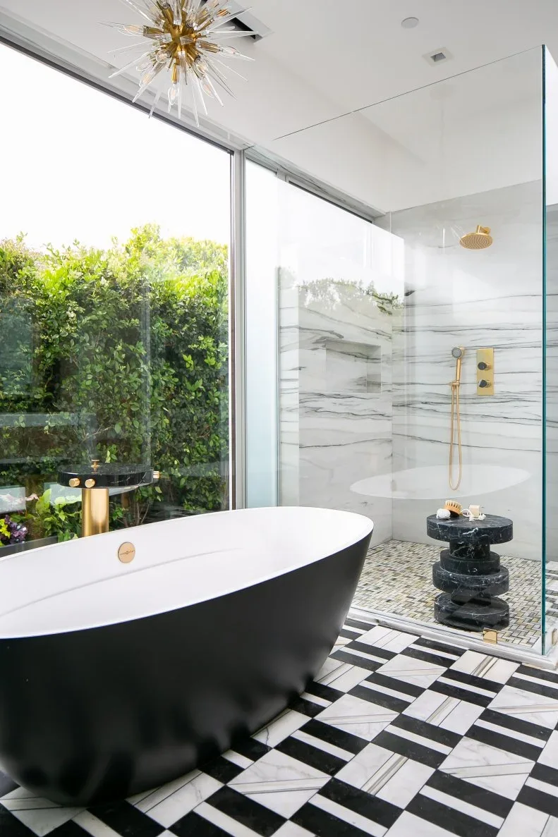 A dark freestanding bath with a white interior sits beside a clear shower screen, pale veined shower walls and a black-and-white patterned floor near broad glazing