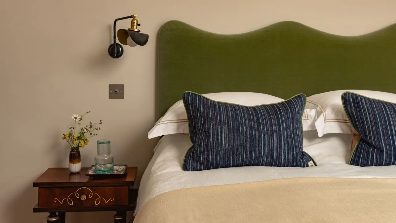 A curved green headboard sits beside a small timber drawer unit and wall-mounted light.
