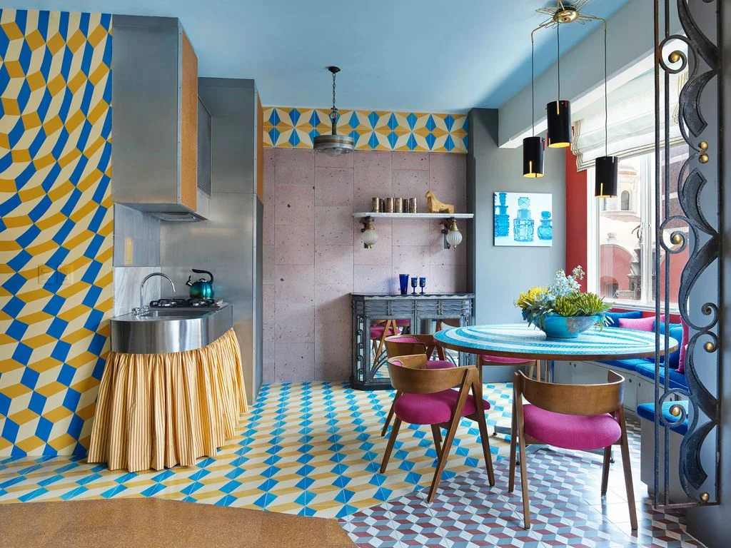 Blue-and-yellow geometric pattern covers a kitchen wall and floor beside a rounded cooking counter with a yellow skirt, pink chairs and a blue circular table