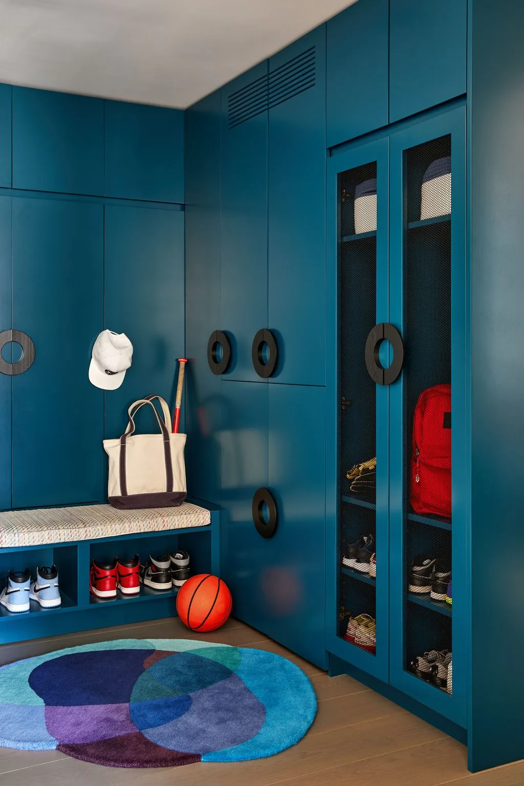 Deep teal mudroom cabinets with large round dark handles, mesh-front shoe compartments, cushioned bench, basketball, baseball bat, and a round multicolor rug.
