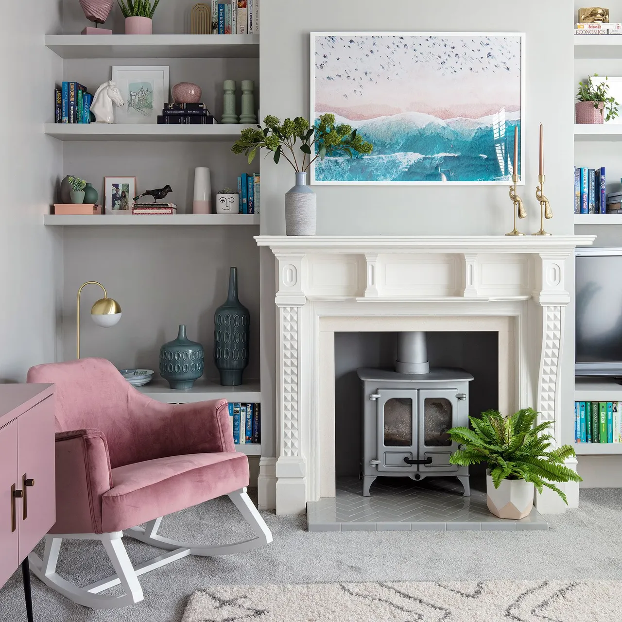 A white mantel sits between light shelves beneath pale artwork beside a pink upholstered chair.