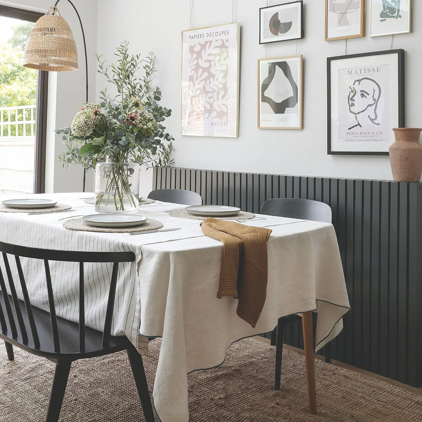 Dining space with a white tablecloth, dark spindle-back chairs, framed wall art, textured rug and an arched floor lamp.