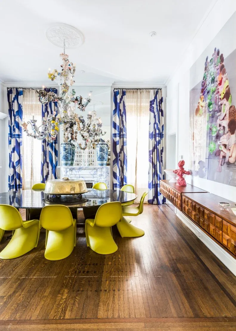 Lime curved chairs around a round glass table beneath an ornate multicolored floral chandelier with blue-and-white curtains.