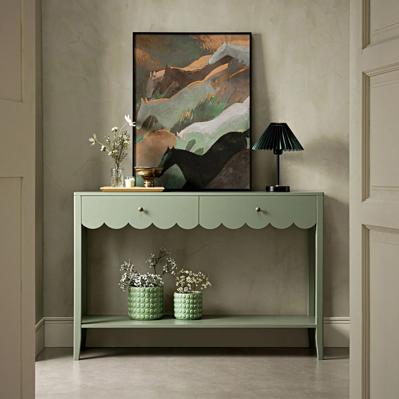 Sage-green scalloped console with black pleated lamp, small green pots and framed abstract picture against a pale wall