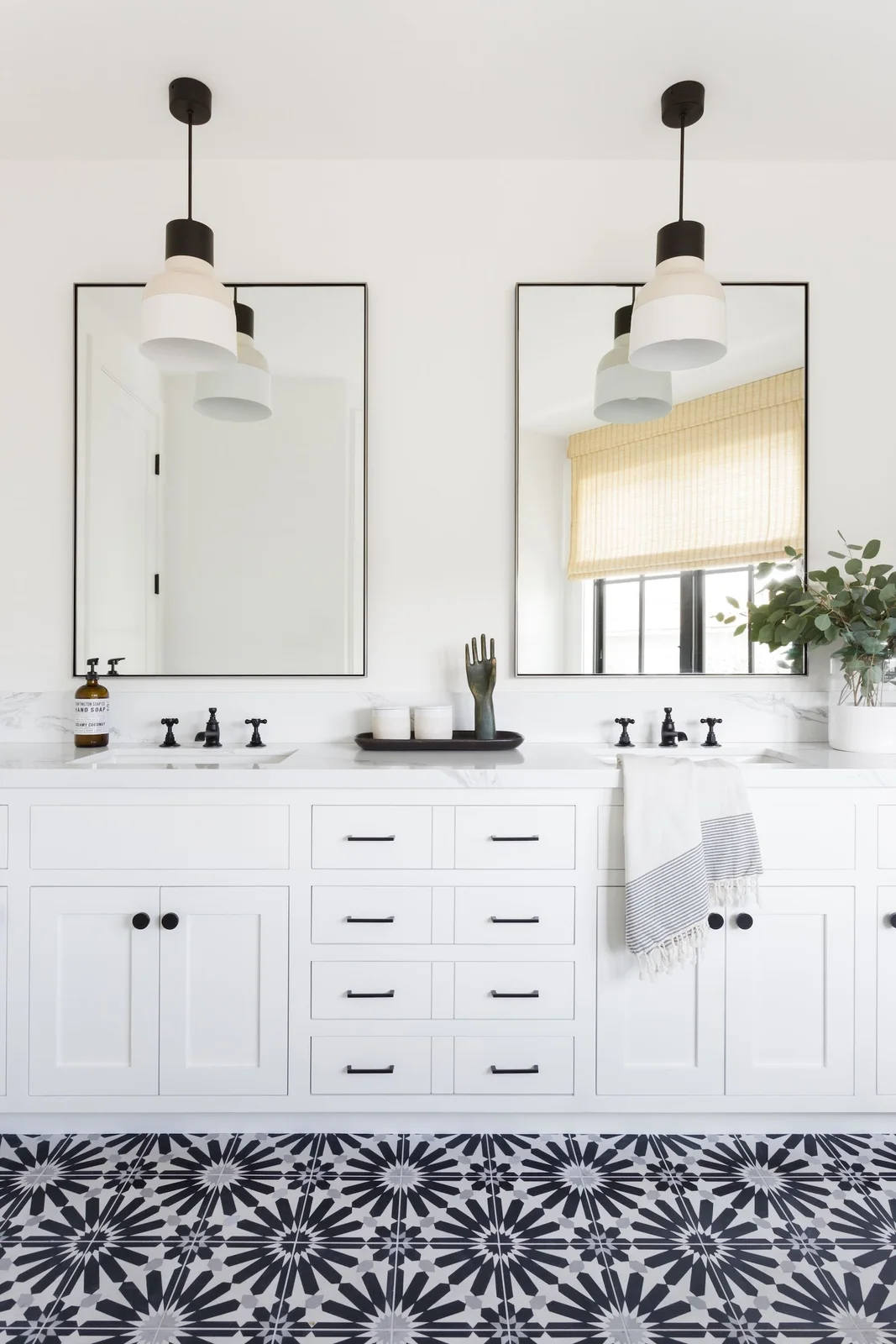 A white double vanity with framed doors and dark handles sits beneath two rectangular mirrors and simple shaded pendants beside a black-and-white patterned floor