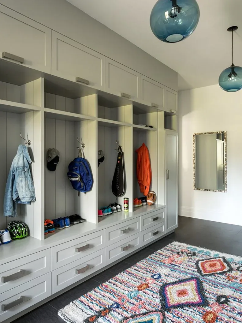 Gray mudroom cubbies and drawers with coats, footwear, blue globe pendants and multicolored textured geometric rug.