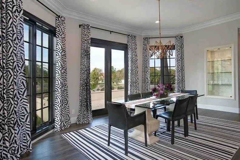 Dark dining chairs around a rectangular table beside black window frames, patterned curtains and a striped rug.