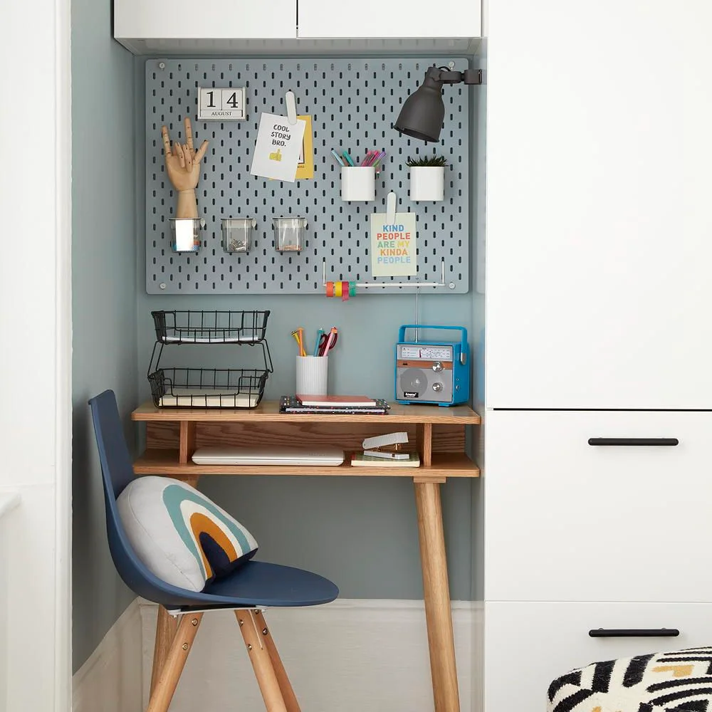 A wood desk sits between white cabinets beneath a gray pegboard with small containers and a dark adjustable lamp beside a blue chair