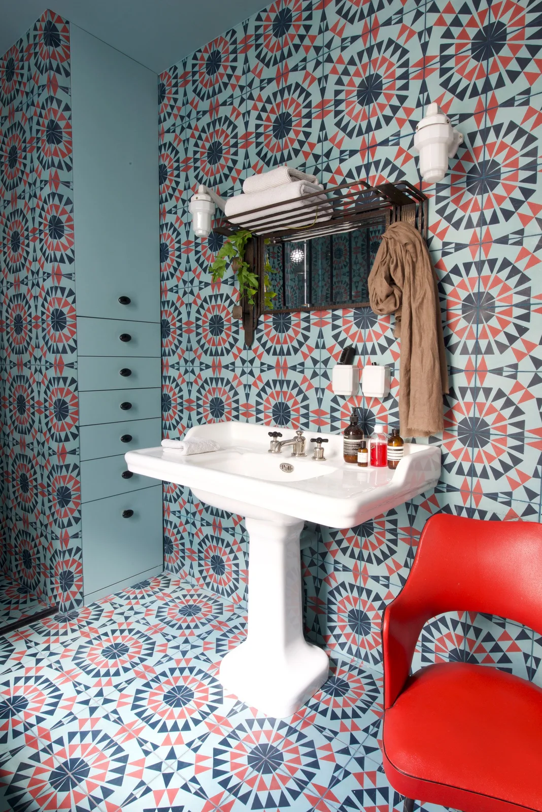 Multicolored geometric tiles cover bathroom walls and floor beside a pale blue cabinet, pedestal basin, wall rack and bright red chair
