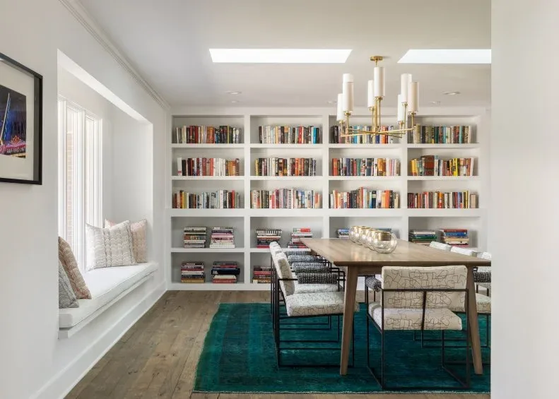 Dining room with a full white bookshelf wall, wood table, patterned chairs, teal rug, window bench, pale walls, and gold-tone chandelier.