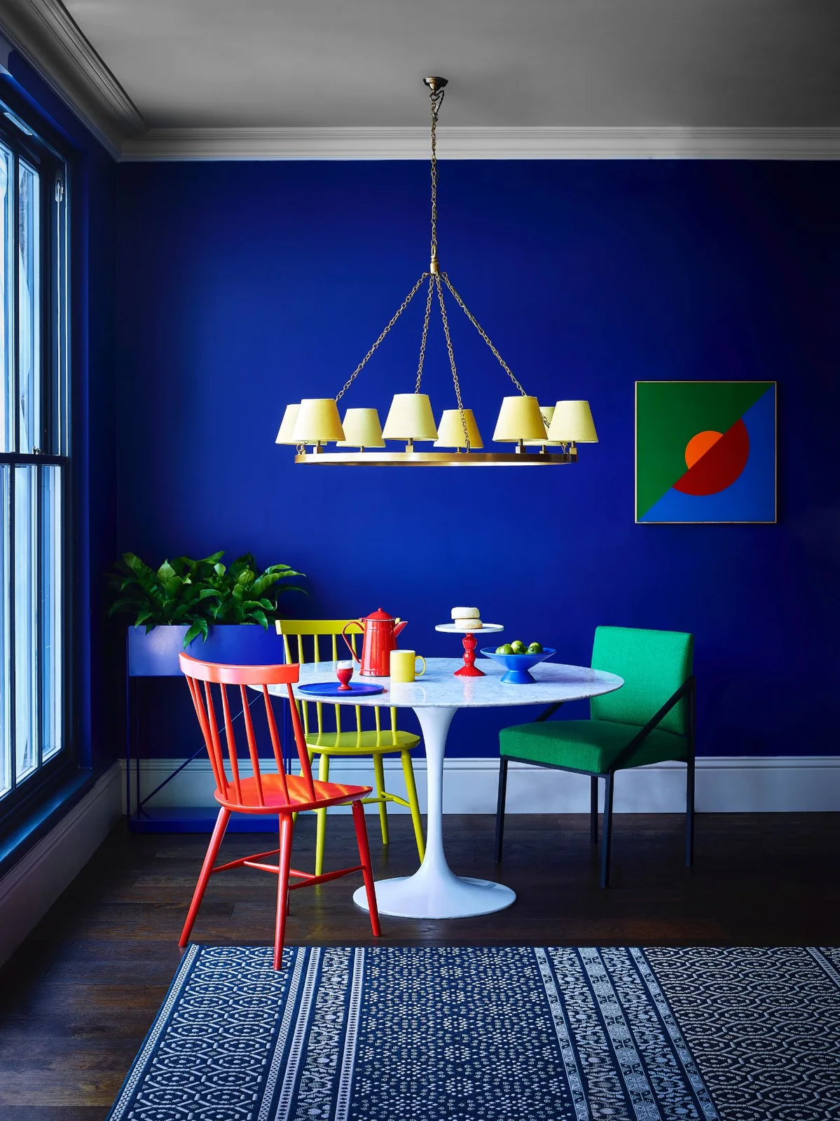 Deep blue walls surround a pale round table with red, yellow and green chairs beneath a yellow-shaded chandelier beside bright geometric artwork