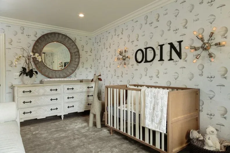 Nursery with natural wood crib, painted dresser, round mirror, illustrated wallpaper and large name letters