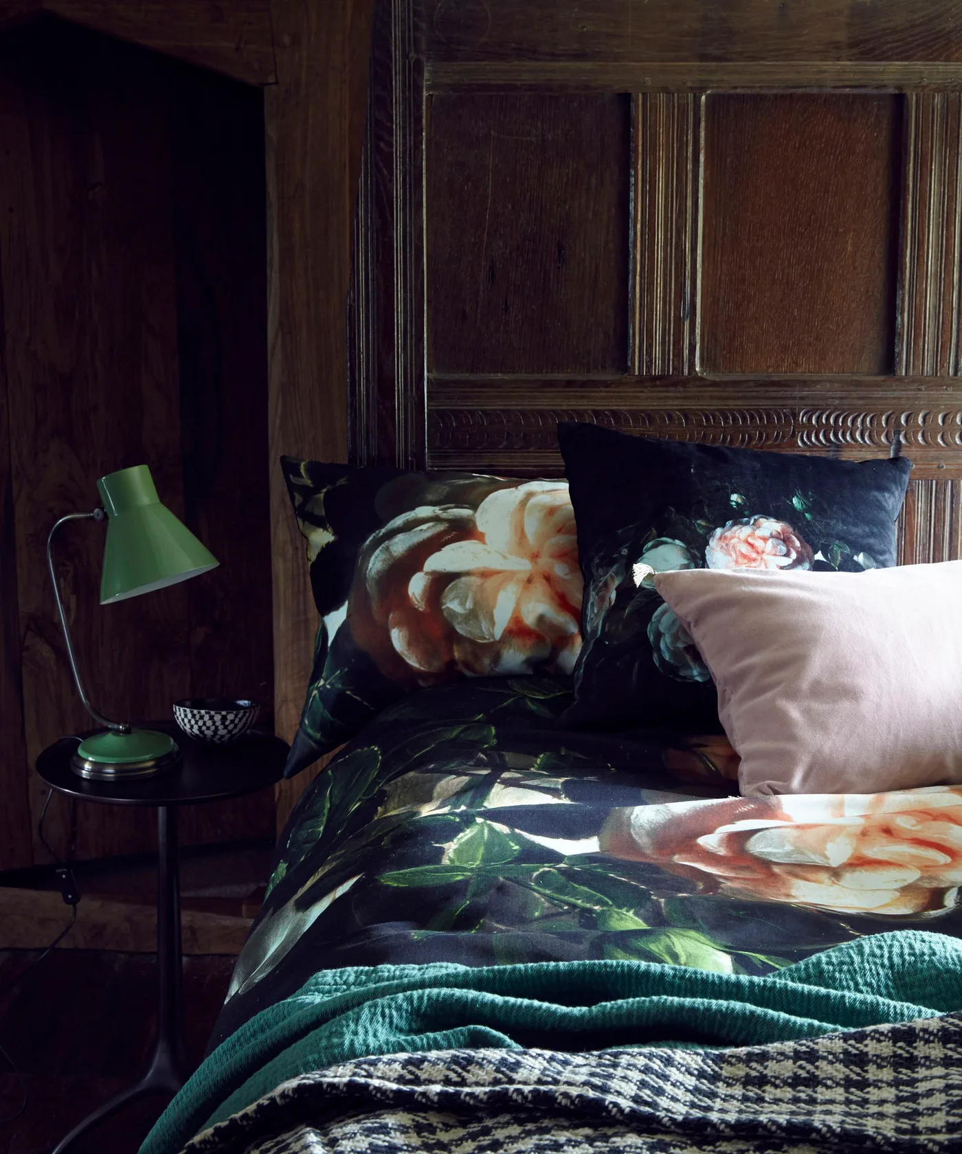 Dark wood wall panels sit behind floral bedding with a pale pink cushion, green throw and adjustable green bedside lamp