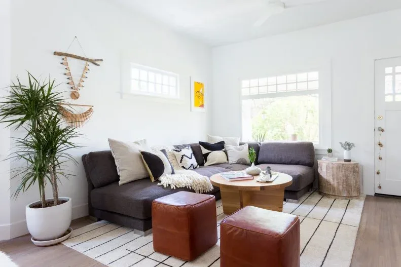 Neutral living room with dark gray sectional, pale grid-patterned rug, round wooden table, two brown square poufs and a tall green plant.
