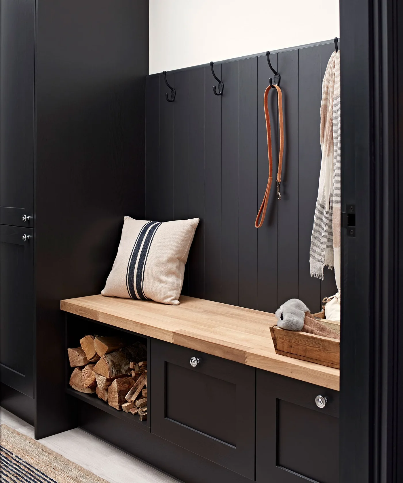 Black paneled mudroom with tall cupboards, coat hooks and an orange leash, wood bench, striped cushion, lower cabinets, and an open firewood compartment.