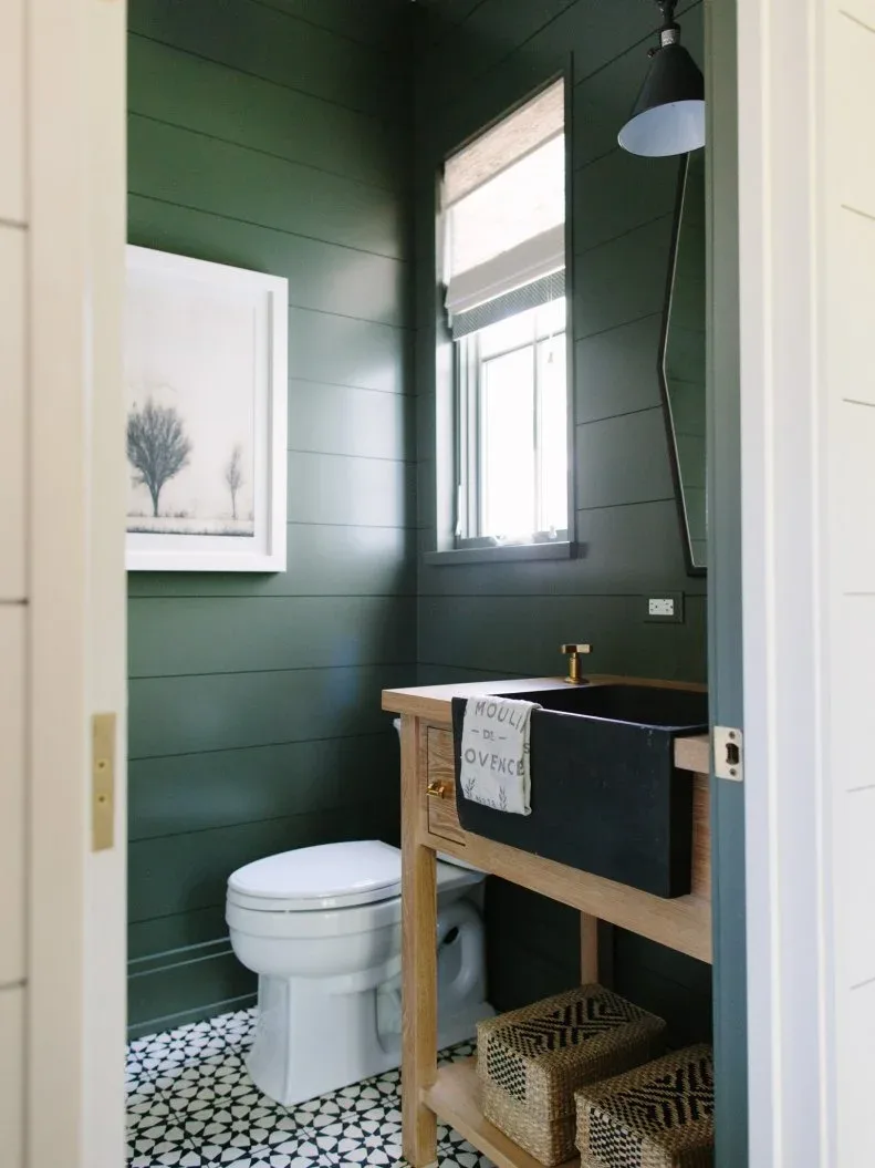Powder room with dark green horizontal walls, pale wooden vanity frame, dark basin, two patterned woven baskets, white toilet and black-and-white tiled floor.