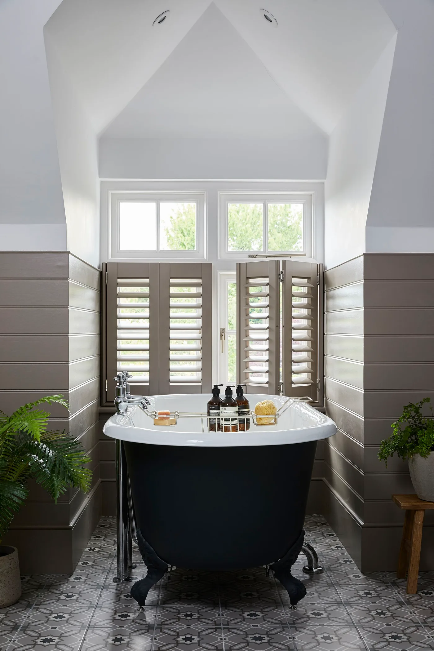 Bathroom with a dark freestanding bath, brown horizontal paneling, patterned floor tiles and lower-window shutters beneath a pale sloping ceiling.