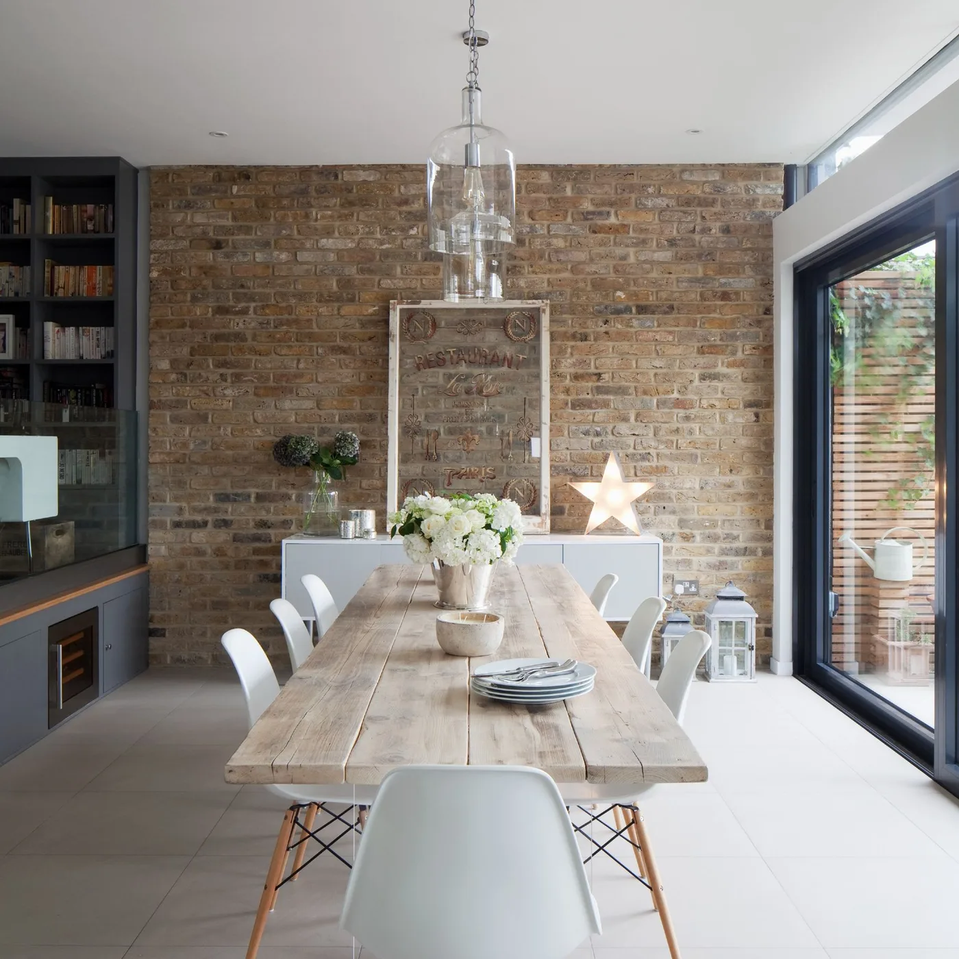 Clear glass pendant above a long pale wood dining table with white chairs and an exposed brick wall