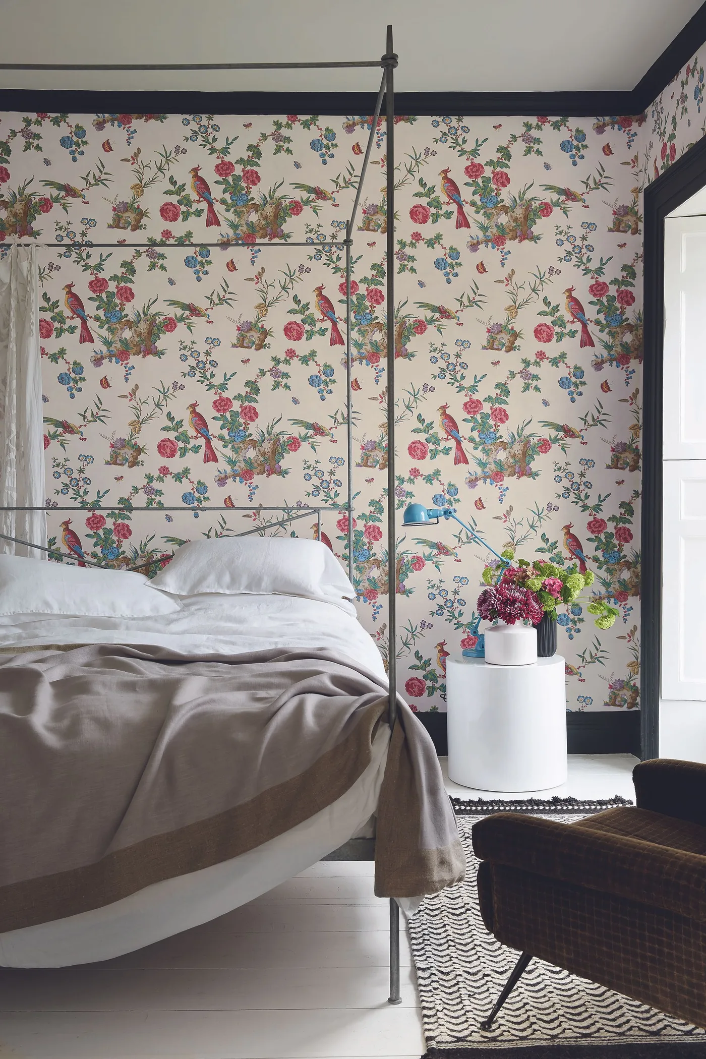 Colorful flowers and birds on pale wallpaper beside a dark four-poster bed and dark ceiling trim.