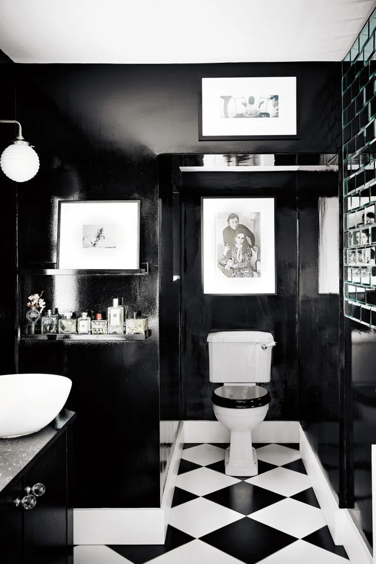A compact washroom has black walls, a white bowl basin, black-and-white diagonal checked flooring and white-framed pictures