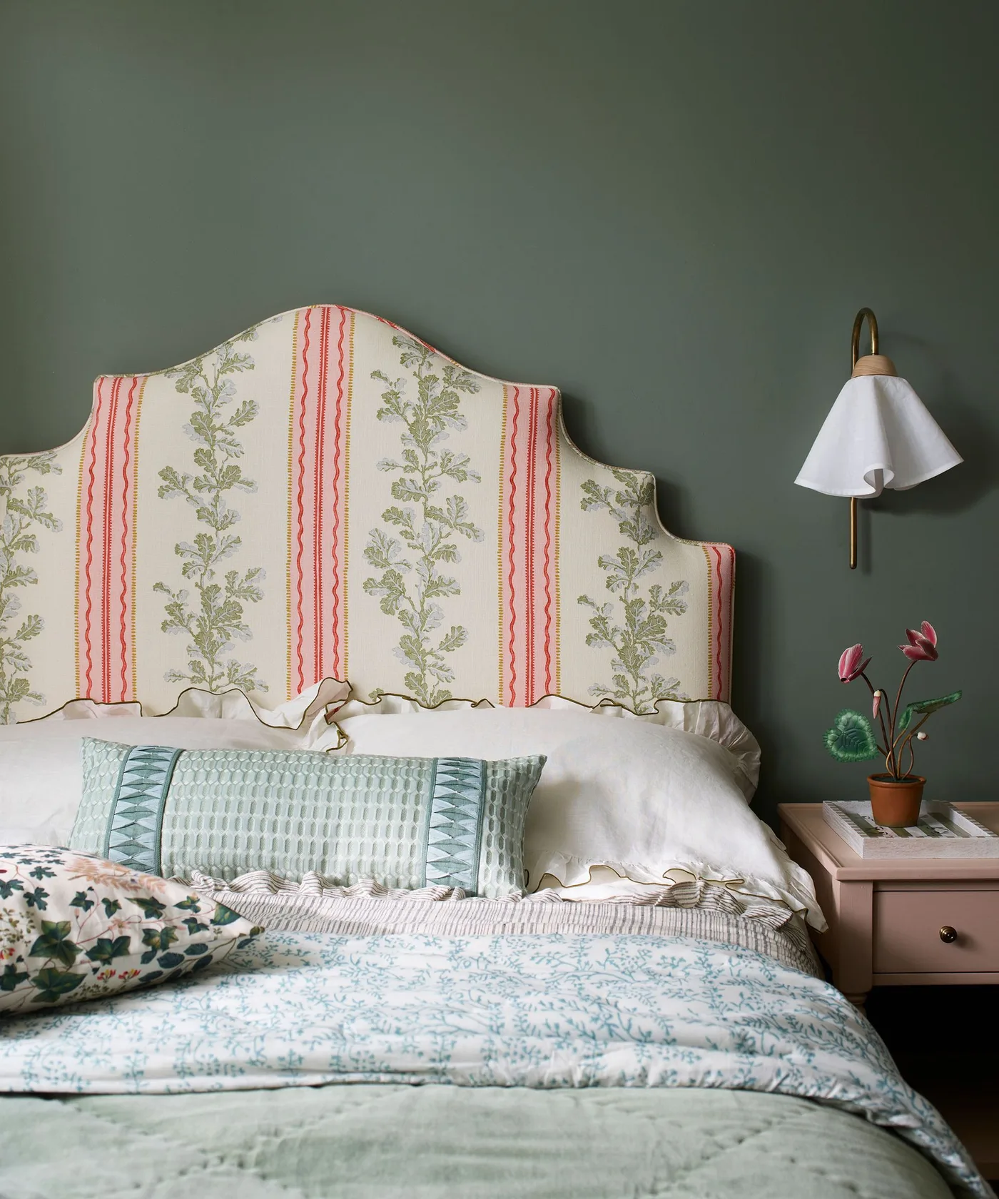 Dark gray-green bedroom wall behind a curved headboard with green and red striped floral upholstery, pale patterned quilt, and wall light.