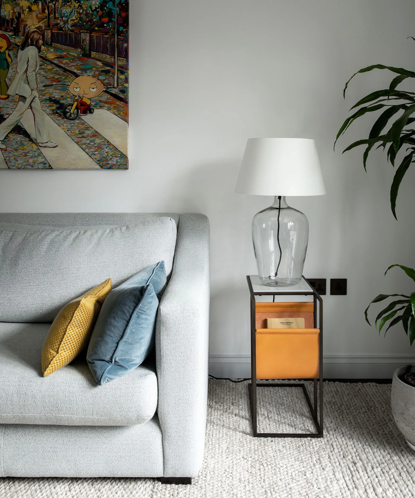 A pale gray sofa with yellow and blue cushions sits beside a narrow dark side unit with warm-colored storage pockets and a clear-base table lamp