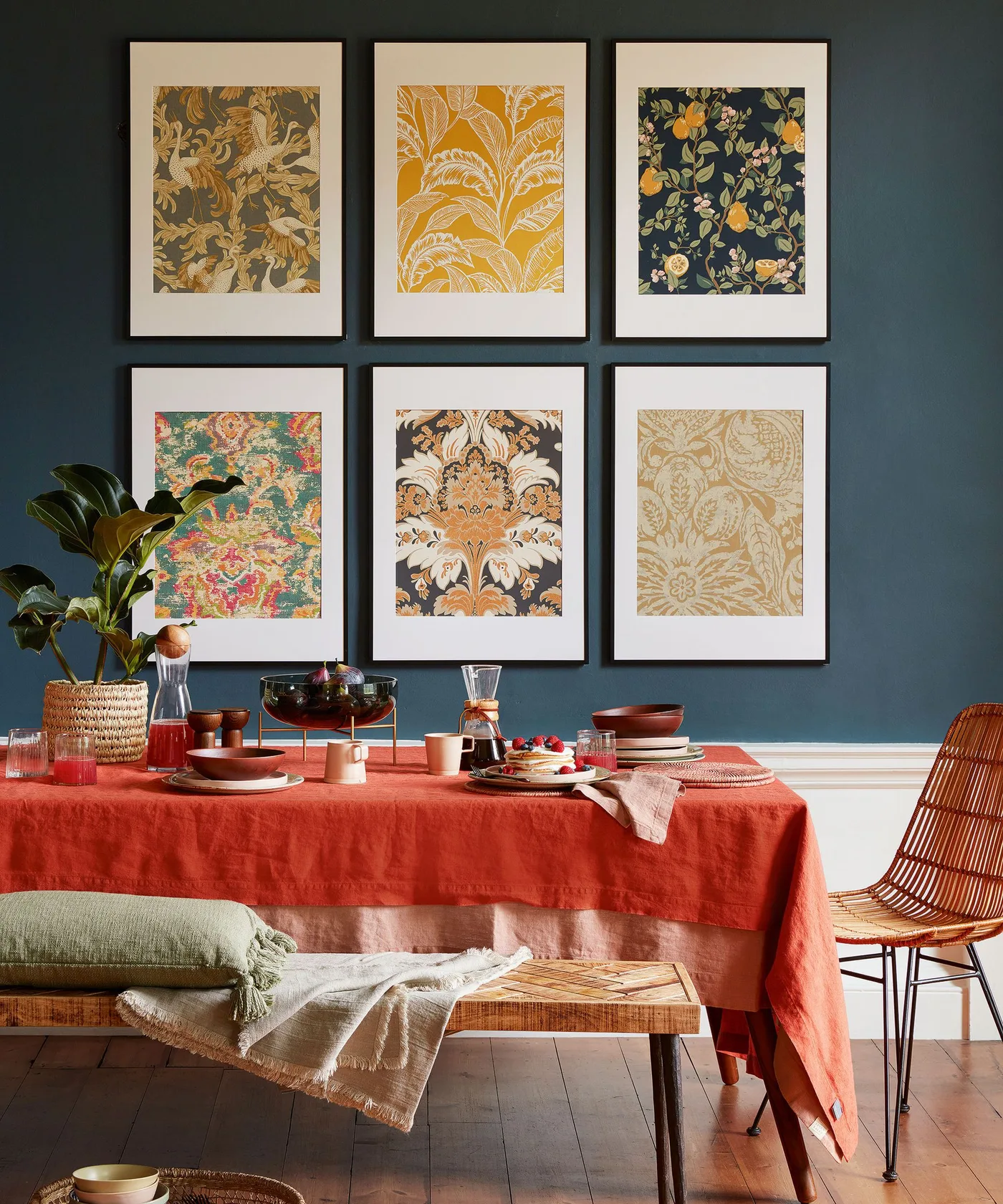 Six patterned prints in matching black frames on a dark blue-green wall, with an orange-red tablecloth, wooden bench, woven chair, and plant.