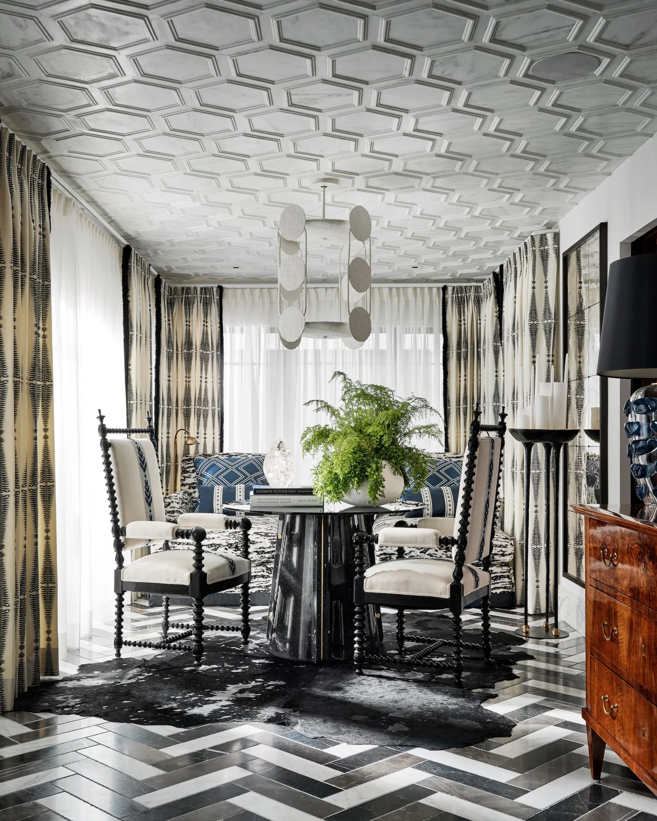 A formal dining room has a black-and-white geometric floor, pale chairs with dark outlines, a dark round table and patterned curtains below a detailed pale ceiling