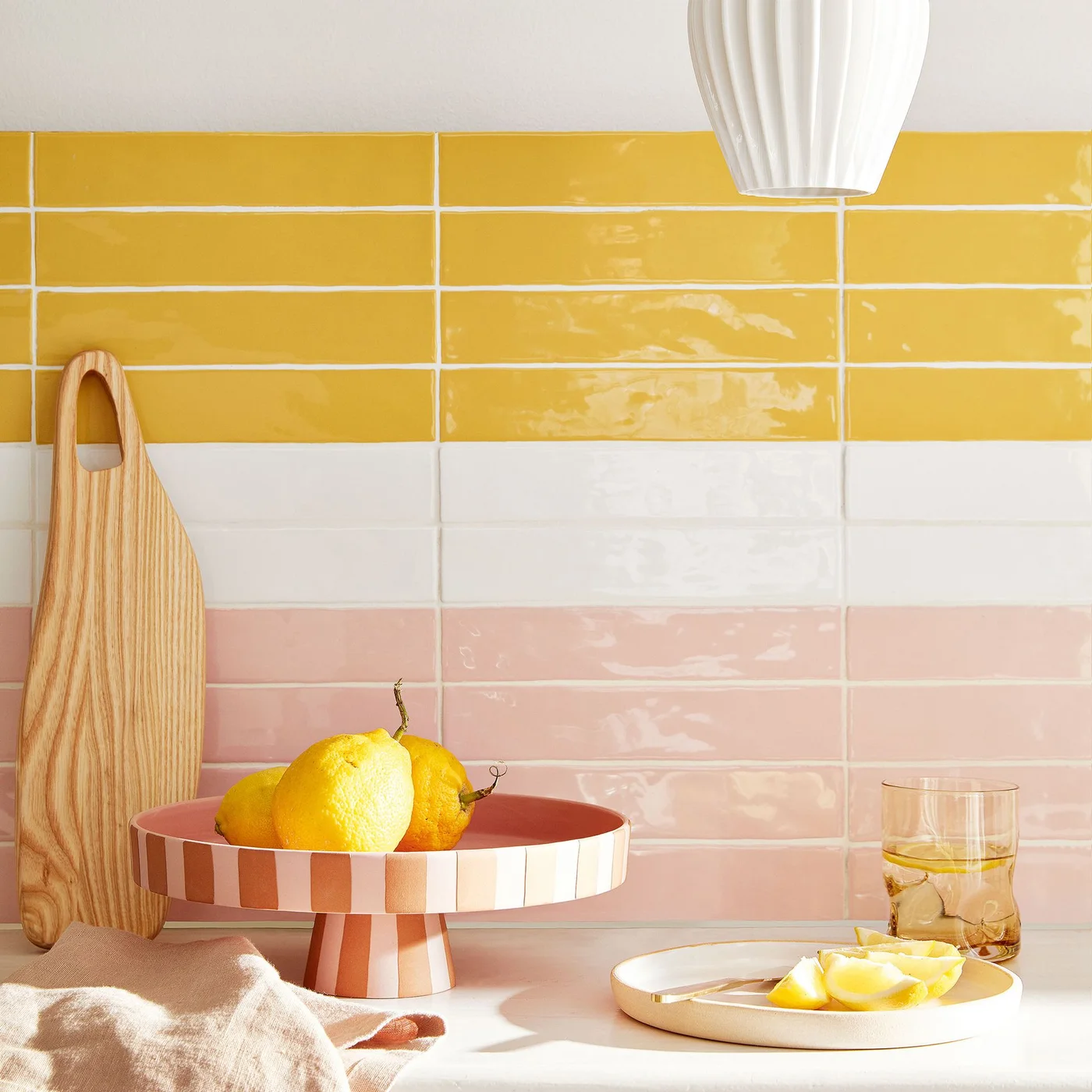 Yellow, white and pale pink rectangular tiles form horizontal bands behind a kitchen counter with wooden boards and a bowl of lemons