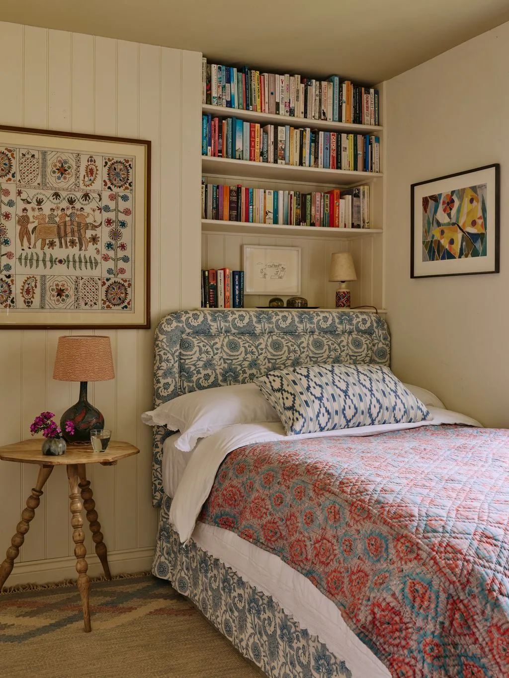 Bedroom with blue patterned upholstered bed, coral quilt, overhead bookshelves, pale paneling, framed artwork and small wood table