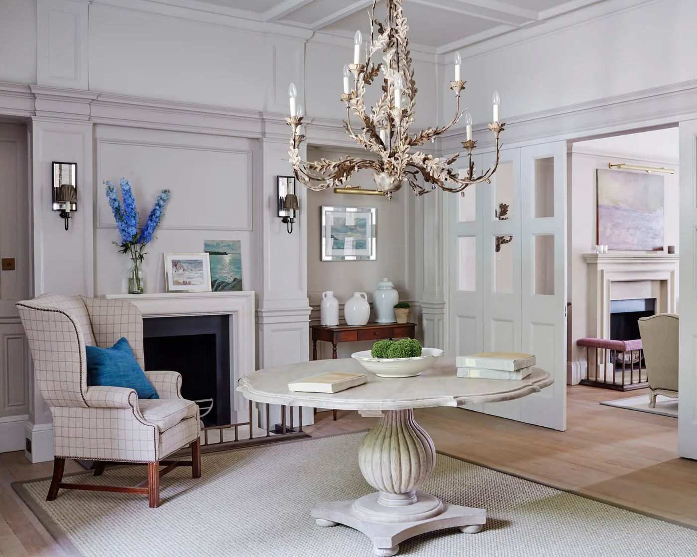 Round pale pedestal table with books and a shallow white bowl in a paneled arrival room with checked chair and chandelier