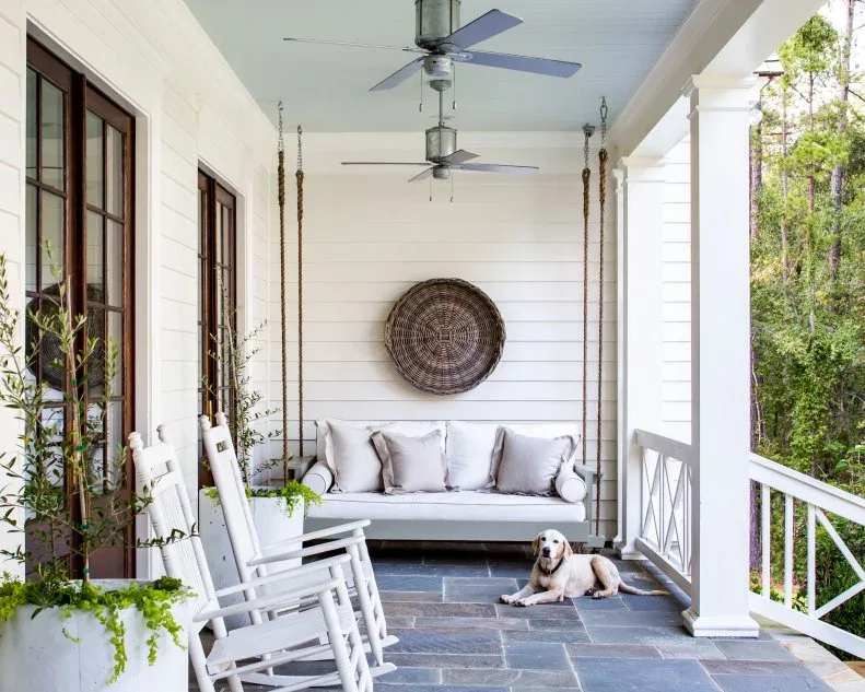White porch swing with gray cushions, white rocking chairs and round woven wall decoration