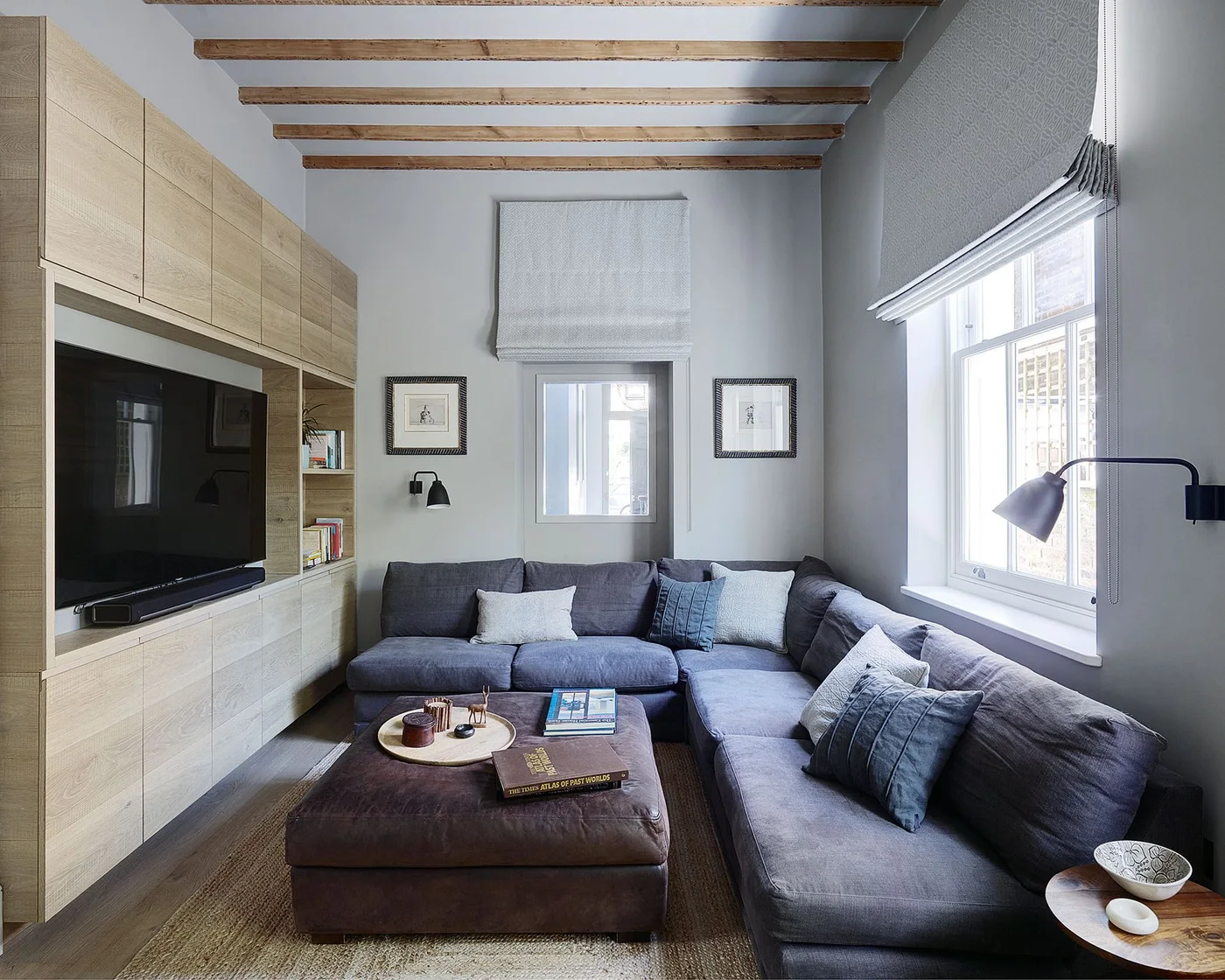 A large television sits within pale wooden wall cabinets opposite a gray corner sofa beneath exposed ceiling beams