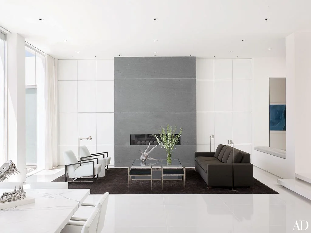 A tall gray fireplace panel interrupts a white paneled wall behind a black sofa, white chairs and a transparent table