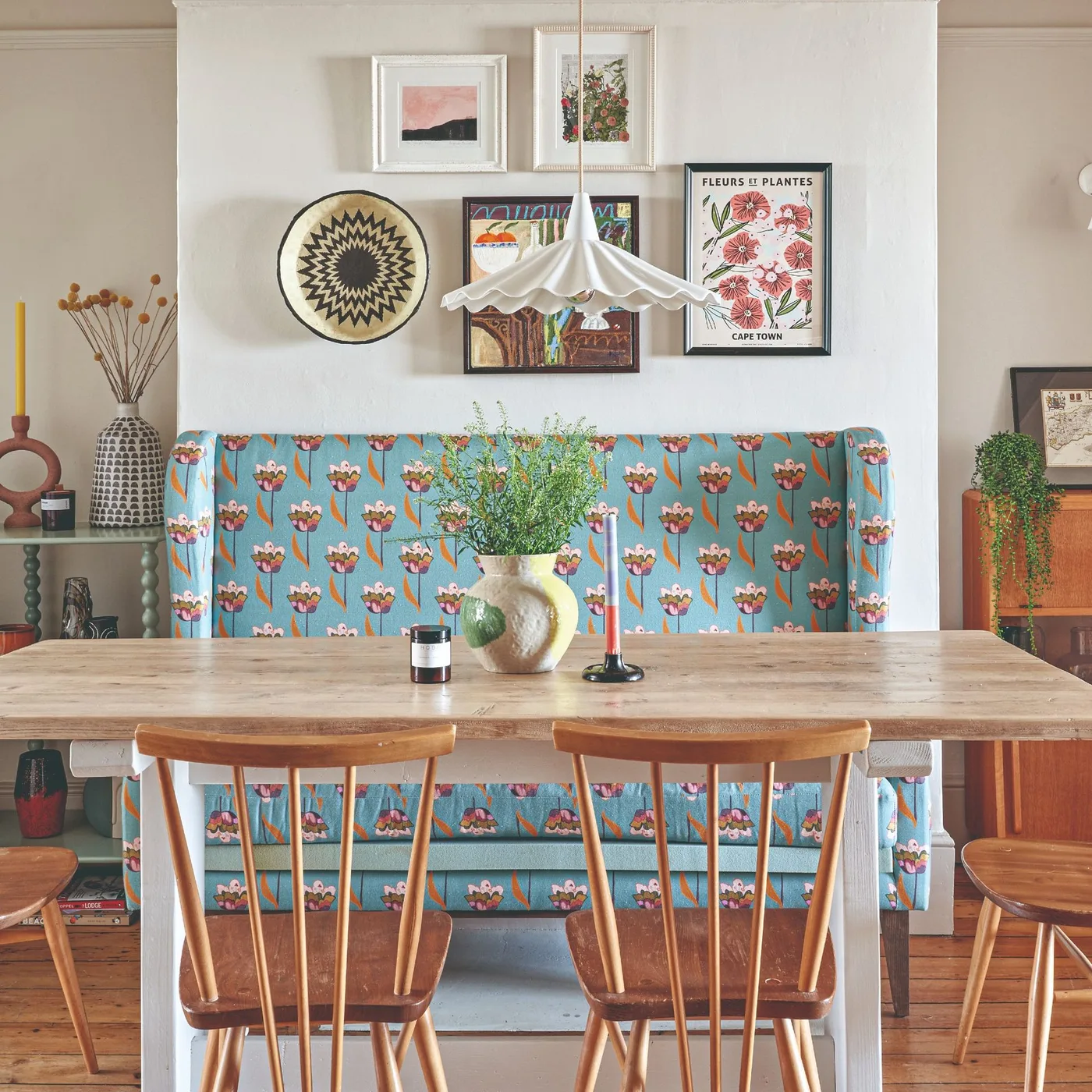 Dining nook with a blue floral upholstered banquette, round wooden table, wooden chairs, framed pictures and a pleated pendant.