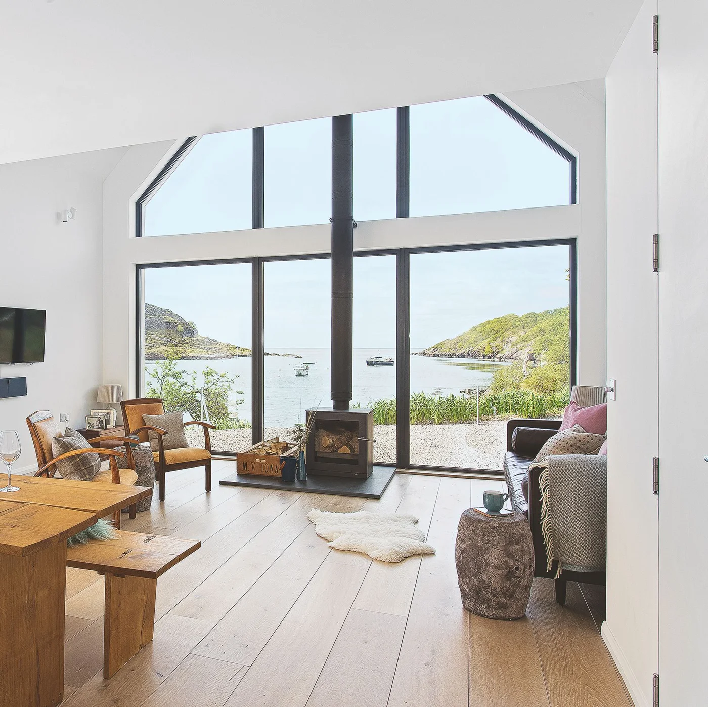 A bright room has a broad waterside window, pale floorboards, wooden furniture and a freestanding dark stove