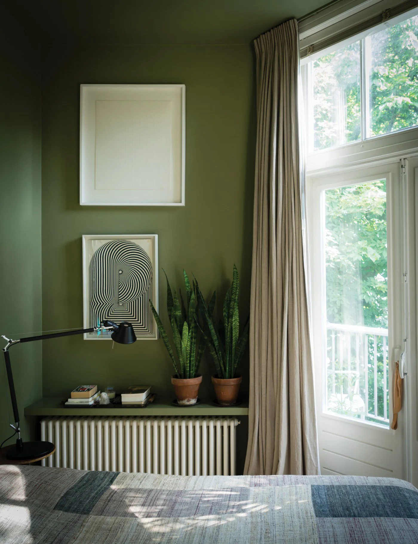 Olive green bedroom walls and ceiling beside a tall bright window, pale curtains, gray striped bedding, abstract prints, and plants.