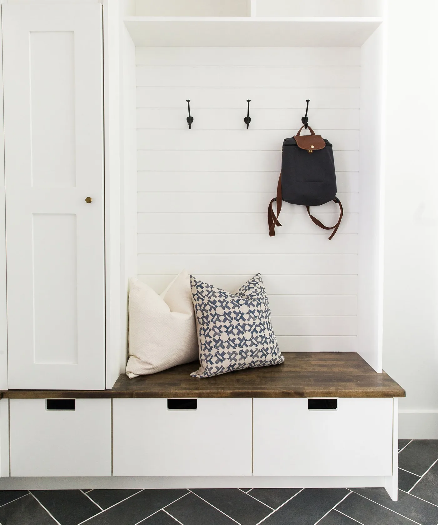 White entry bench with three lower drawers, a dark wood seat, black coat hooks and two patterned cushions