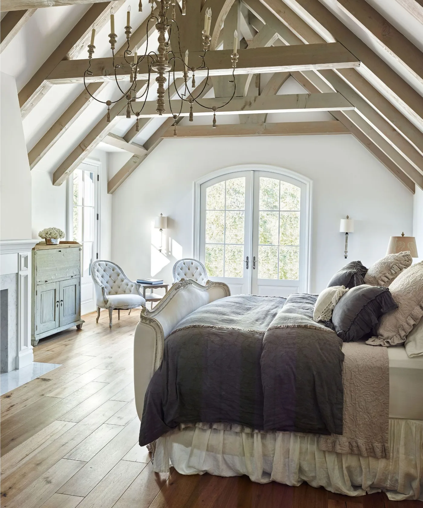 Country bedroom with exposed angled roof beams, a pale shaped bed, dark gray coverlet, two chairs and glazed double doors.
