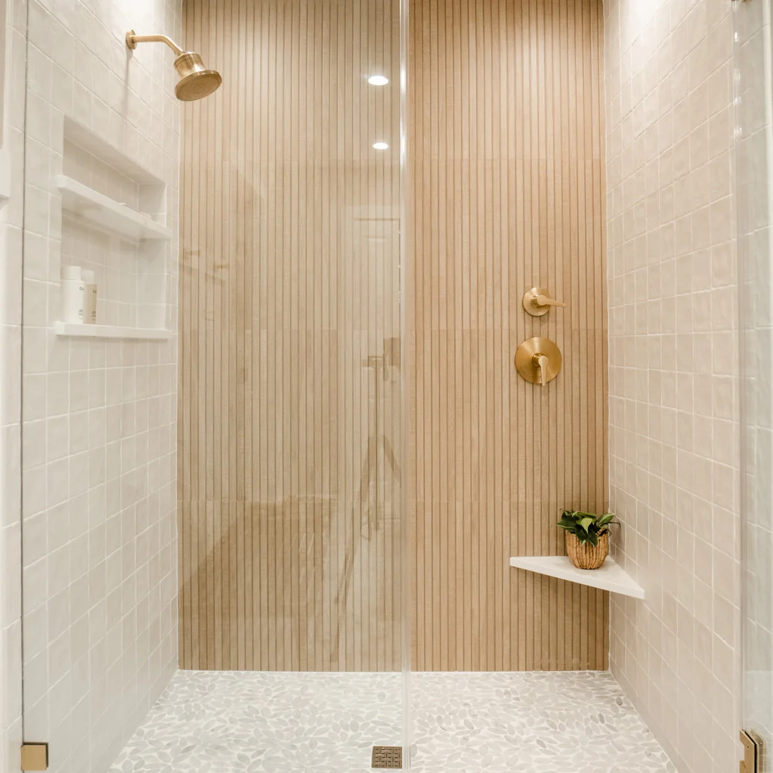 Narrow wood-look vertical strips form a shower feature wall beside pale rectangular tiles, brass-colored fittings and a corner shelf