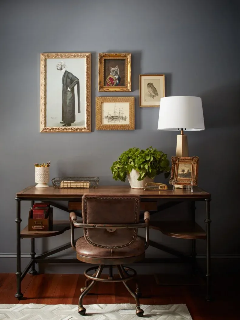 Wood and dark-framed writing desk with brown swivel chair, gold-toned framed gallery, pale lamp and dark gray wall