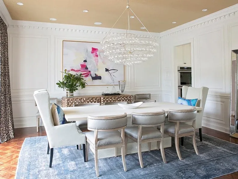 Wide clear tiered chandelier above a pale dining table and upholstered chairs, with paneled white walls, colorful abstract artwork, and a blue rug.