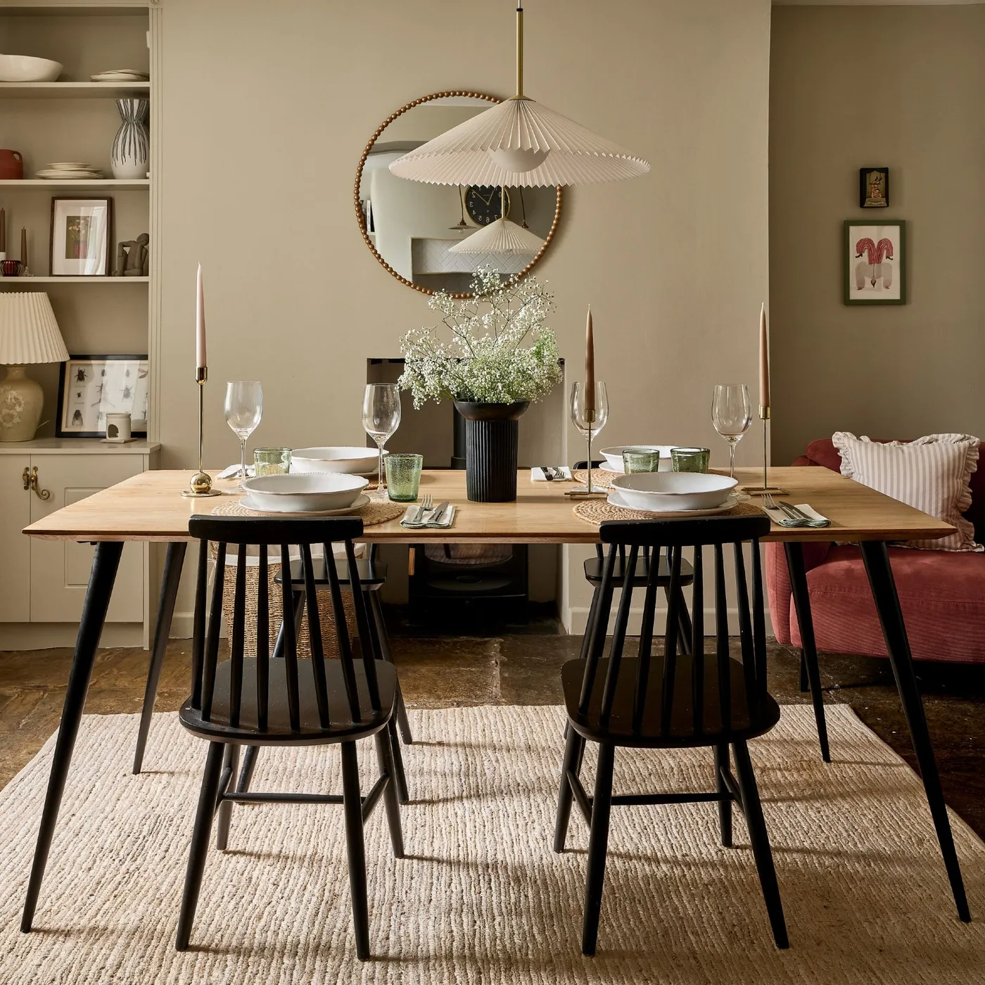 Black spindle chairs sit around a wooden table beneath a pale pleated pendant beside warm neutral walls and a round mirror