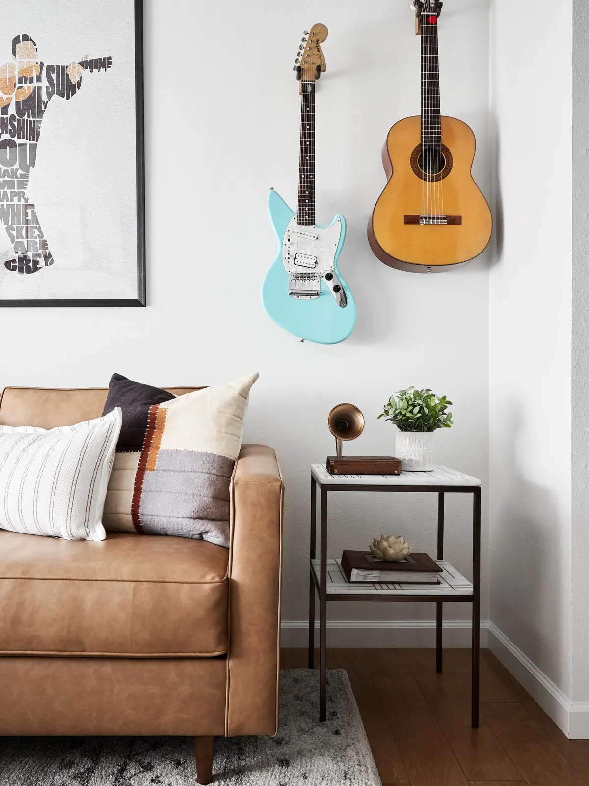 Guitars hang above a tan sofa beside a small pale side table.